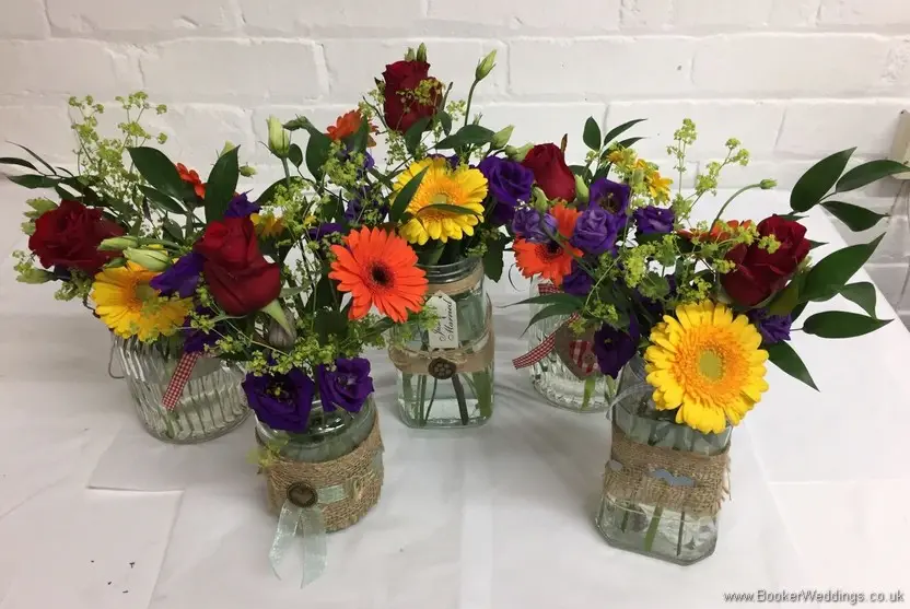 Bright and vibrant jam jar table centrepieces for the reception featuring roses, gerberas, lissianthus and alchamelia molis side view
