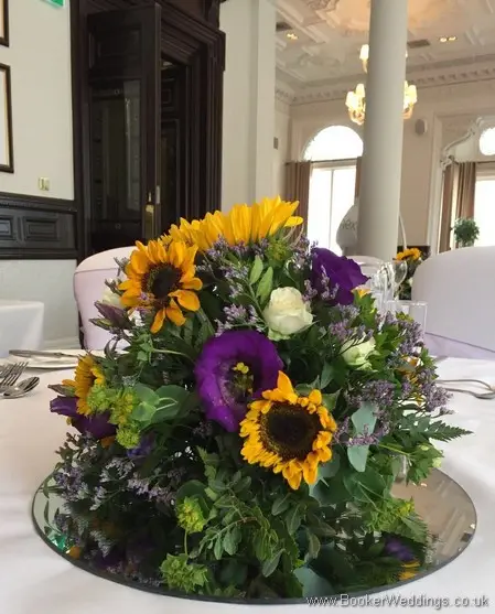 Low posy table arrangements on mirror plate with sunflowers, yellow gerberas, white spray roses, purple lissianthus at Doubletree by Hilton Liverpool